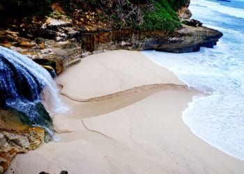 Tebing karang yang membingkai pantai dengan ombak biru jernih dan pasir lembut—suasana alami sekaligus menyejukkan. (Sumber: Indonesiakaya.com)